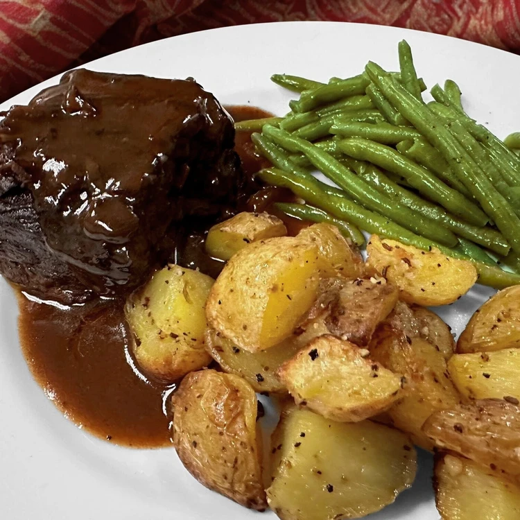 A plate with pot roast topped with brown gravy, roasted potatoes, and green beans.