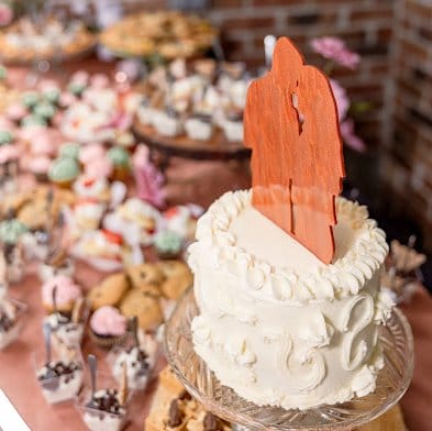 A white frosted cake with a silhouette topper of a couple stands on a dessert table filled with assorted sweets and pastries.