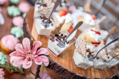 Mini dessert cups with chocolate chips and wafers are arranged on a wooden stand, with pink artificial flowers in the foreground.