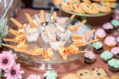 A glass cake stand holds mini desserts in cups with spoons, piped cream pastries, cookies, and frosted cupcakes on a table with pink flowers.